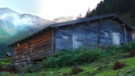 Old-wooden-alpine-hut-close-up-after-sunset-with-light-rays-behind-and-an-alpine-landscape-with-trees-and-alpine-mountains-in-the-background-and-green-bushes-in-front