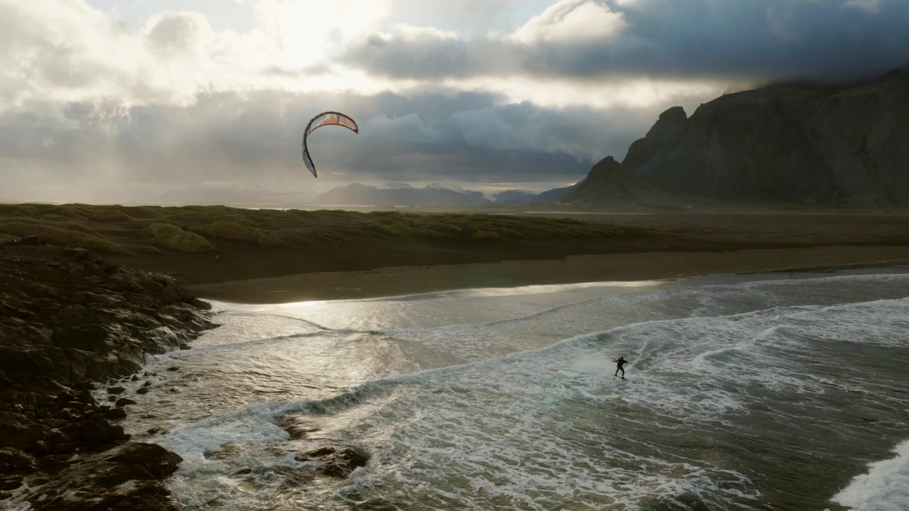 Kitesurfer tracks along black volcanic beach beneath steep Reynisfjall cliffs and breaking Atlantic surf, South Coast of Iceland, drone pullback tracking.