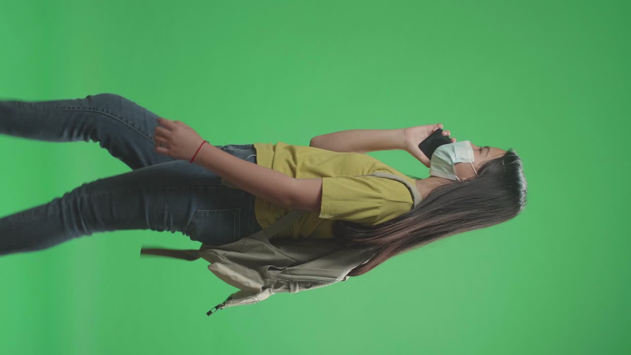 vista lateral de una estudiante asiática con una máscara y hablando en el teléfono móvil mientras camina a la escuela en pantalla verde clave de croma