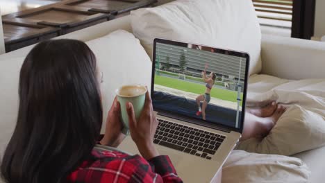 compuesto de una mujer sentada en casa sosteniendo un café viendo un evento de salto de altura en un portátil