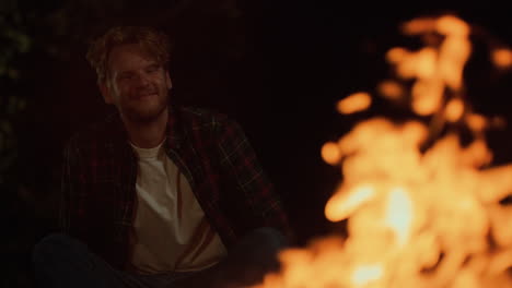 un campista sonriente de cerca disfruta de una fogata en una noche oscura. un hombre alegre se sienta en el bosque.