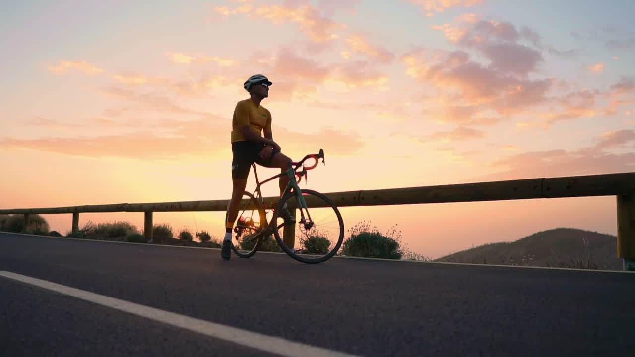 Sportsman in yellow t-shirt and gear rests on bike atop mountain, admiring mountains and sunset following training