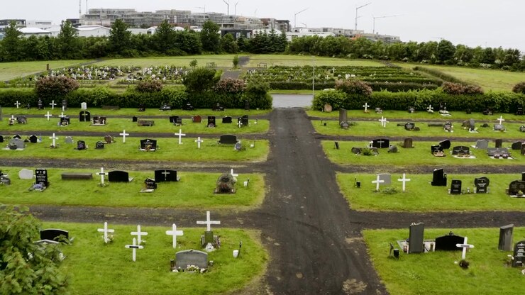 Vista dall'alto di un cimitero verde lussureggiante con numerose lapidi e croci bianche. Una città moderna con edifici in costruzione è visibile sullo sfondo sotto un cielo coperto