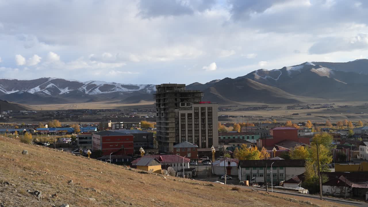 A right-panning, high-angle view of Uliastai, Mongolia, on a cool autumn day. Smoke rises from a chimney over the city, with majestic, snow-capped mountains in the distant background