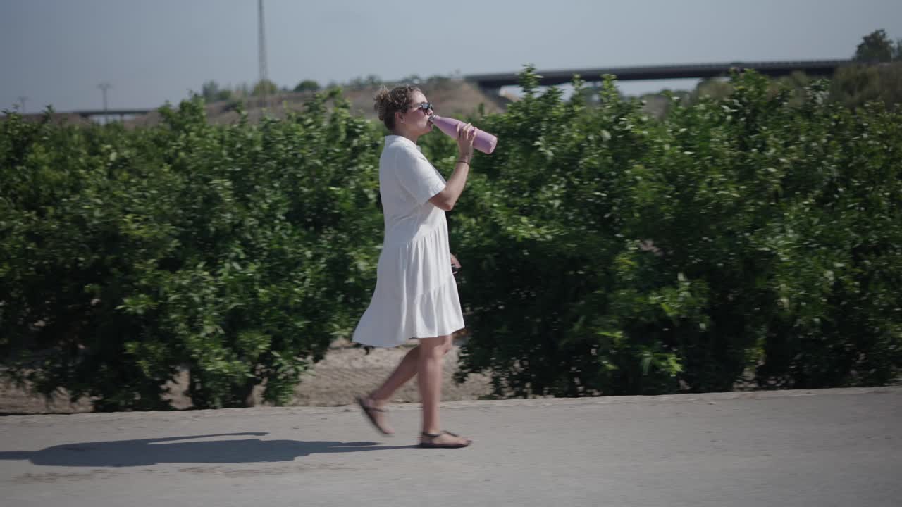 Woman Walking in Citrus Grove with Pink Bottle