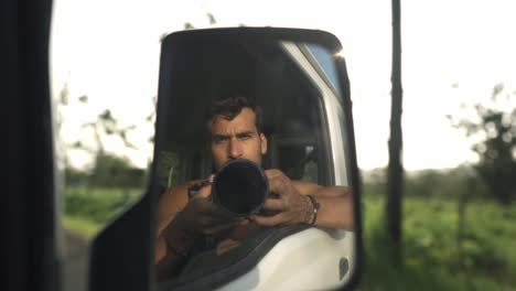 a man takes a selfie reflected in the side mirror of a car, with a camera obscuring his face. the background shows the lush, green landscape of la fortuna, costa rica