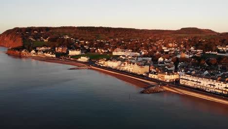 Antenne-über-Dem-Ärmelkanal-Mit-Blick-Auf-Die-Esplanade-Der-Stadt-Sidmouth,-Die-In-Goldenes-Sonnenaufgangslicht-Getaucht-Ist