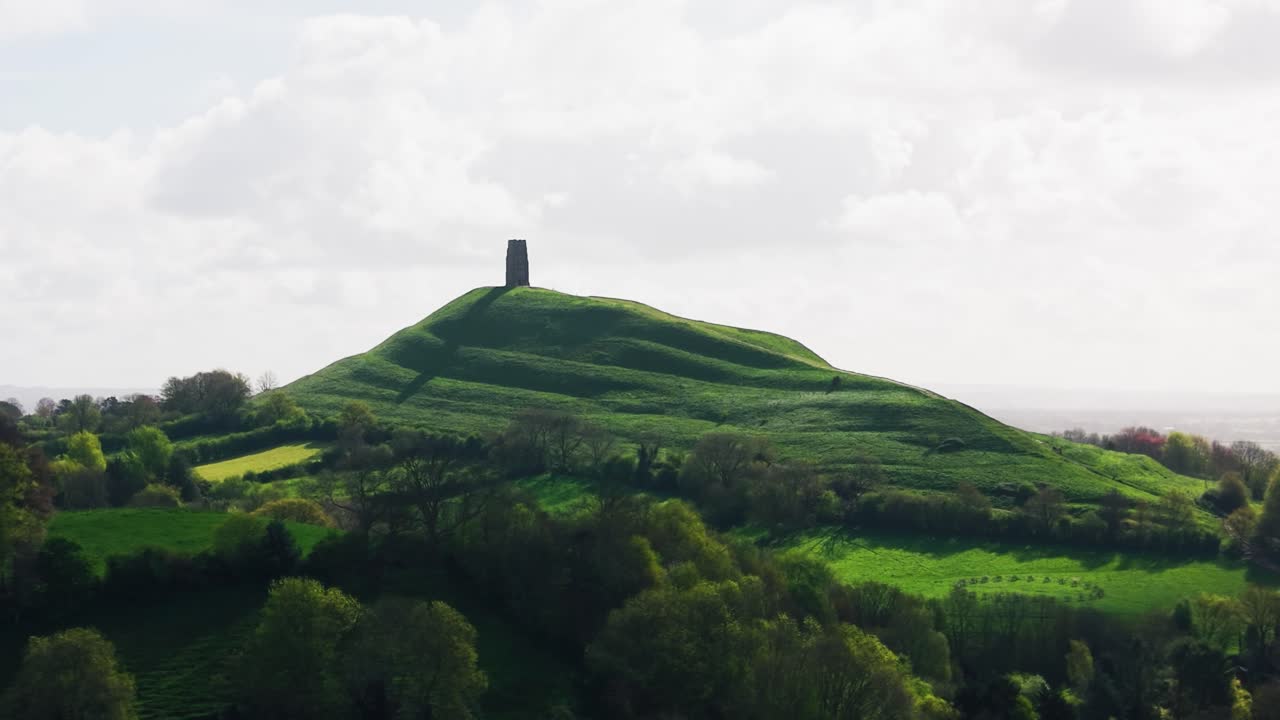 Luftfoto af Glastonbury Tor med det historiske tårn på toppen af en grøn bakke omgivet af et bølgende landskab i Somerset, England, Storbritannien