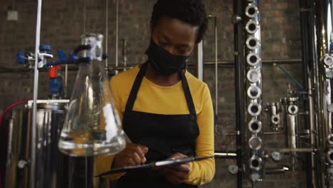 African-american-woman-in-face-mask-working-at-distillery-checking-equipment,-writing-on-clipboard