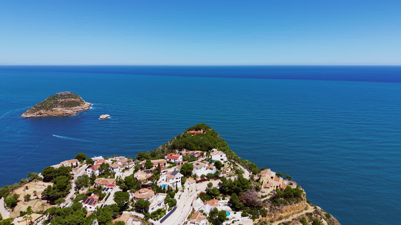 Village méditerranéen perché sur une colline avec des villas aux toits de tuiles en terre cuite nichées parmi des arbres verdoyants, surplombant une mer d'un bleu profond, une petite île rocheuse et un bateau de passage visible en arrière-plan
