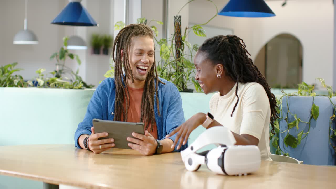 Two coworkers sitting at wood table holding tablet near VR headset gesturing and laughing at screen