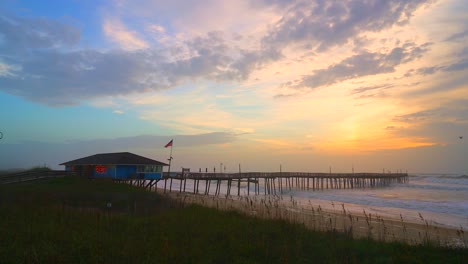 Video-Eines-Majestätischen-Und-Farbenfrohen-Sonnenaufgangs-Am-Avalon-Pier-Mit-Einer-Amerikanischen-Flagge,-Die-In-Der-Ferne-Weht