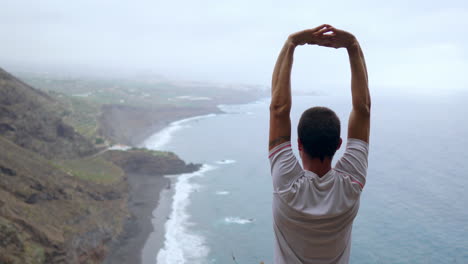 En-Lo-Alto-De-Un-Acantilado,-Un-Hombre-Levanta-Las-Manos-E-Inhala-La-Brisa-Del-Océano-Mientras-Practica-Yoga,-Abrazando-La-Atmósfera-Relajante.