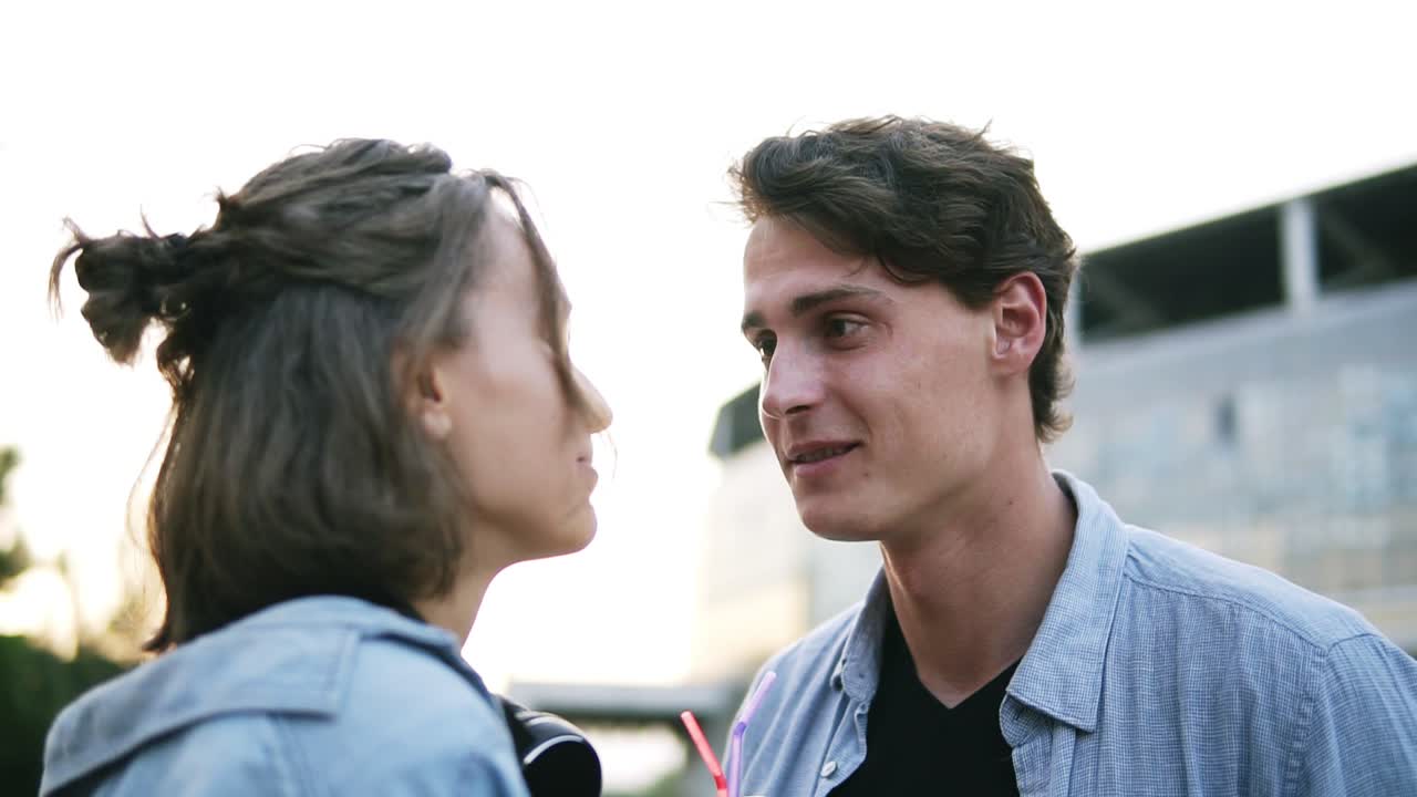 젊은 매력적인 부부는 함께 플라스틱 컵에서 술을 마시고 있습니다. 공원에서 서서 부드럽게 키스하고 있습니다. 느린 움직임