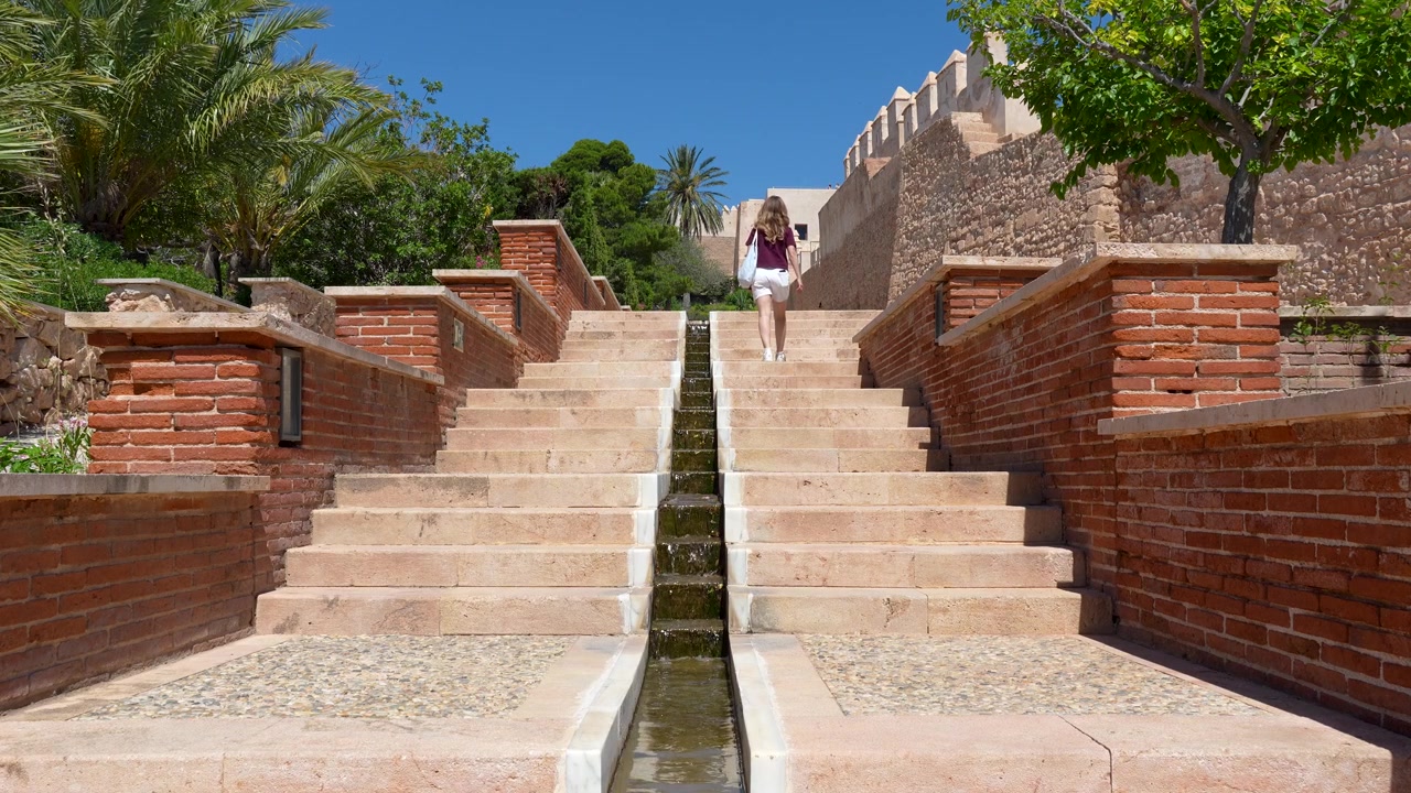 Almeria, İspanya'daki Alcazaba'nın içinde, tarihi kale duvarları ve bahçelerle çevrili taş merdivenlerde yürüyen genç kadın