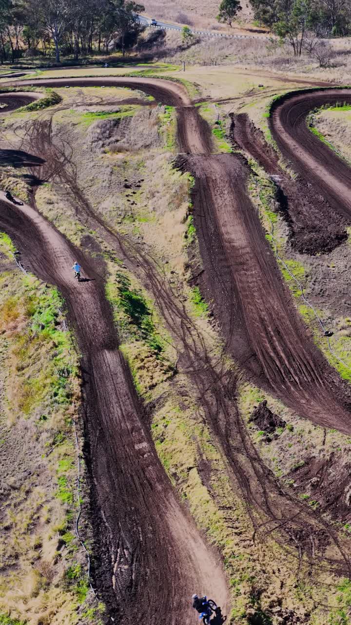 Un pilota di moto da cross accelera in curva e compie un salto su una tortuosa pista da motocross all'aperto, ripreso da un drone ad alta angolazione in pieno giorno.