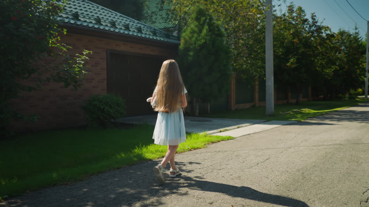 Girl in blue gown with net fabric walks slowly down calm residential street under bright sun casting soft shadow on ground with brick house, lush greenery, and roadside drainage visible in background