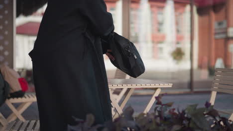 woman sits at a cafe, places her bag on the table, and unzips it, revealing her thigh, with an urban background featuring buildings and a nearby bag