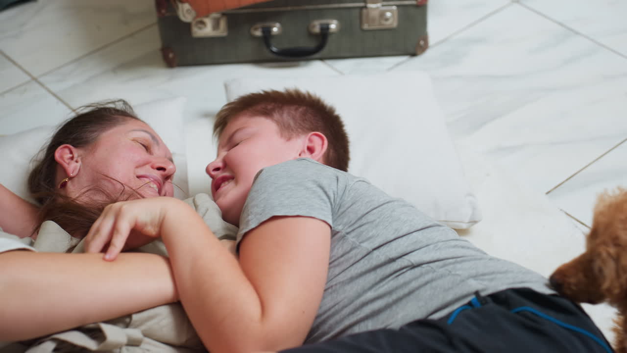 mom and son lying on white pillows laughing and playing together cozy bonding moment boy smiling eyes closed mother smiling warmly marble tile floor background joyful home family interaction