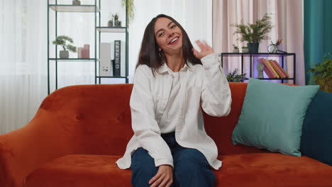 Young-woman-smiling-friendly-at-camera,-waving-hands-gesturing-hello,-hi,-greeting-at-home-on-couch