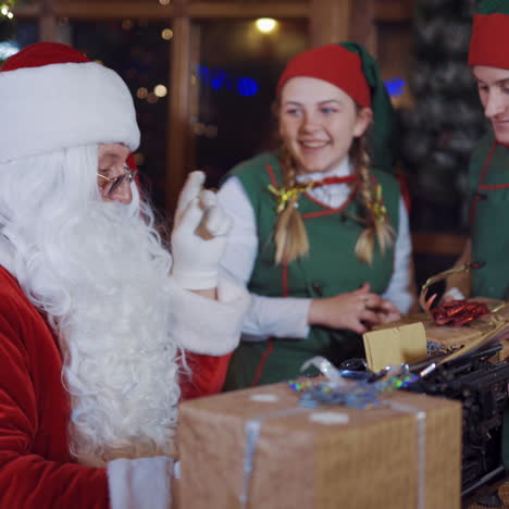 santa claus and two elves with presents. happy elves in fairy costumes are talking to santa near christmas tree indoors.