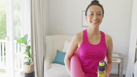 Retrato-De-Una-Mujer-Asiática-Feliz-Sosteniendo-Una-Estera-De-Yoga-Y-Una-Botella-De-Agua-En-El-Dormitorio,-En-Cámara-Lenta