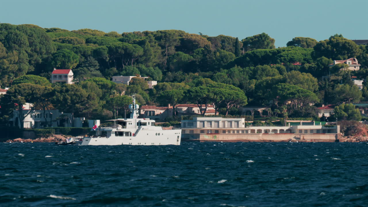 Luxury white yacht sailing near Mediterranean coastline with elegant villas surrounded by trees