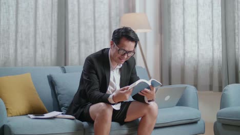 hombre de negocios asiático en chaqueta y pantalones cortos leyendo un libro después de trabajar con una computadora portátil en casa