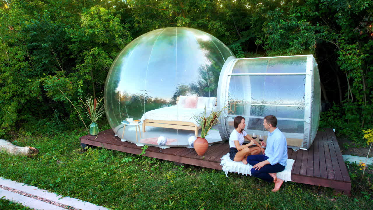Vista aerea di una coppia felice che beve vino vicino a una tenda a bolla trasparente in un glamping. Natura circostante