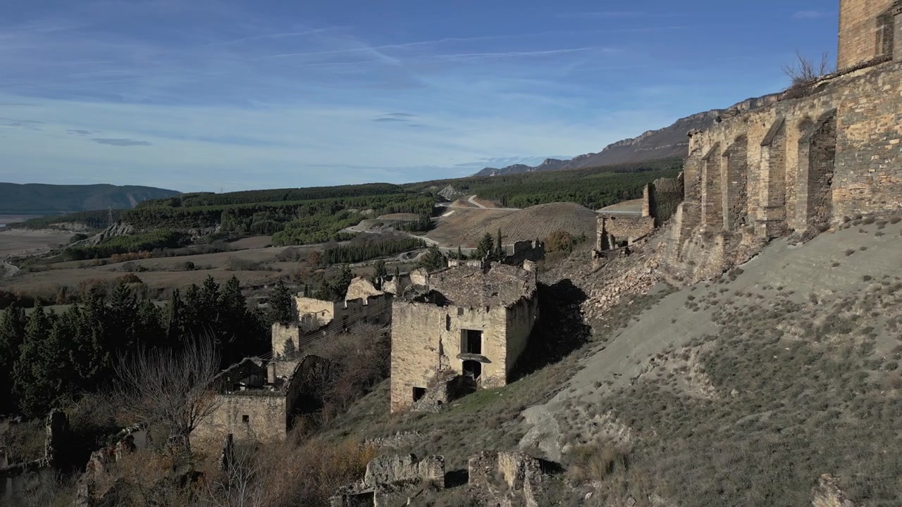 Panoramica drammatica che mostra la città fantasma abbandonata di Esco in Aragona, Spagna, con le sue case in pietra in rovina che crollano su un fianco di collina sotto un cielo azzurro limpido in una giornata di sole.