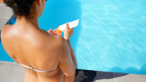 High-angle-view-of-young-mixed-race-woman-applying-sunscreen-on-her-shoulder-near-pool-4k