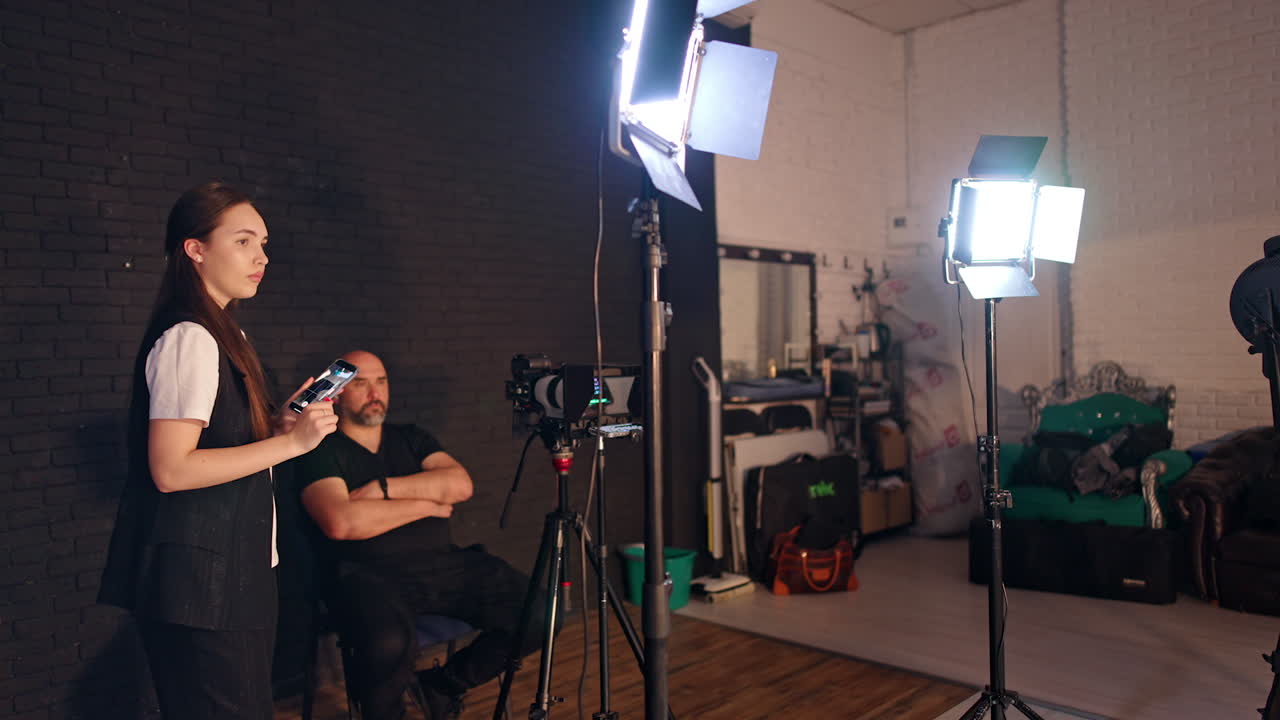 Assisting girl fixes the filming equipment and moves back to the wall starting to take a video on her phone. Cameraman sitting on the chair looks ahead. Studio footage backstage.
