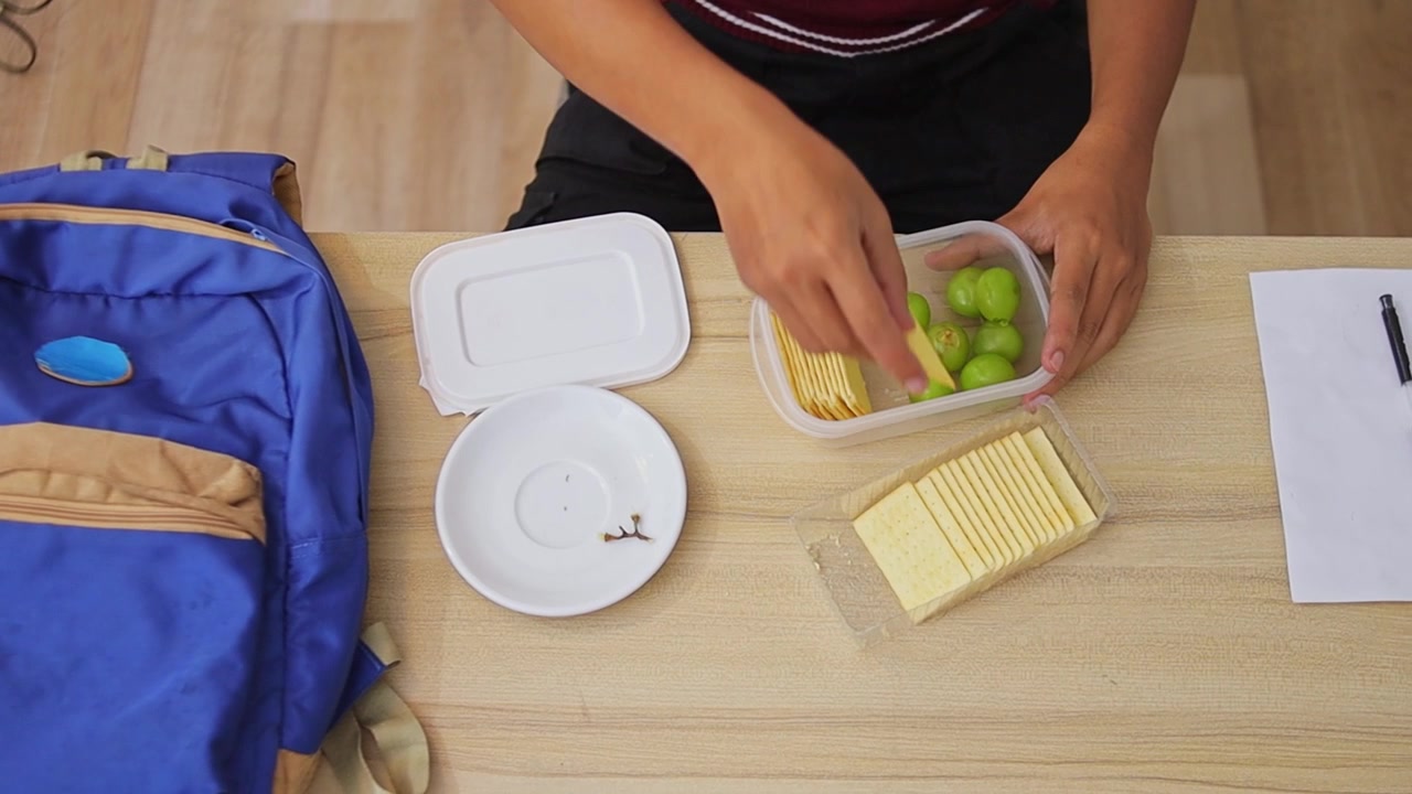 Toma cenital de un estudiante guardando galletas saladas de queso en una lonchera junto a uvas verdes.