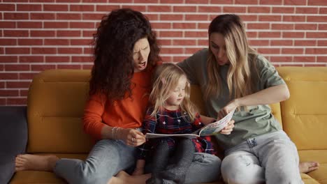 two women read a book together to a small preschool girl sitting on the sofa at home