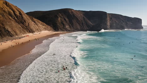 An-aerial-drone-shot-of-wave-hitting-a-beach-next-to-a-mountain-range-where-people-are-swiming-and-surfing-in-Algarve,-Portugal