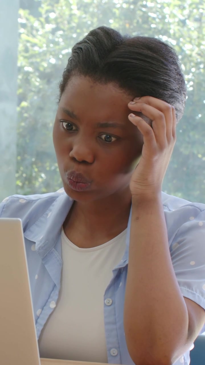 Vertical video: Reacting to laptop alert, woman touching head at desk by window, blue shirt, ring