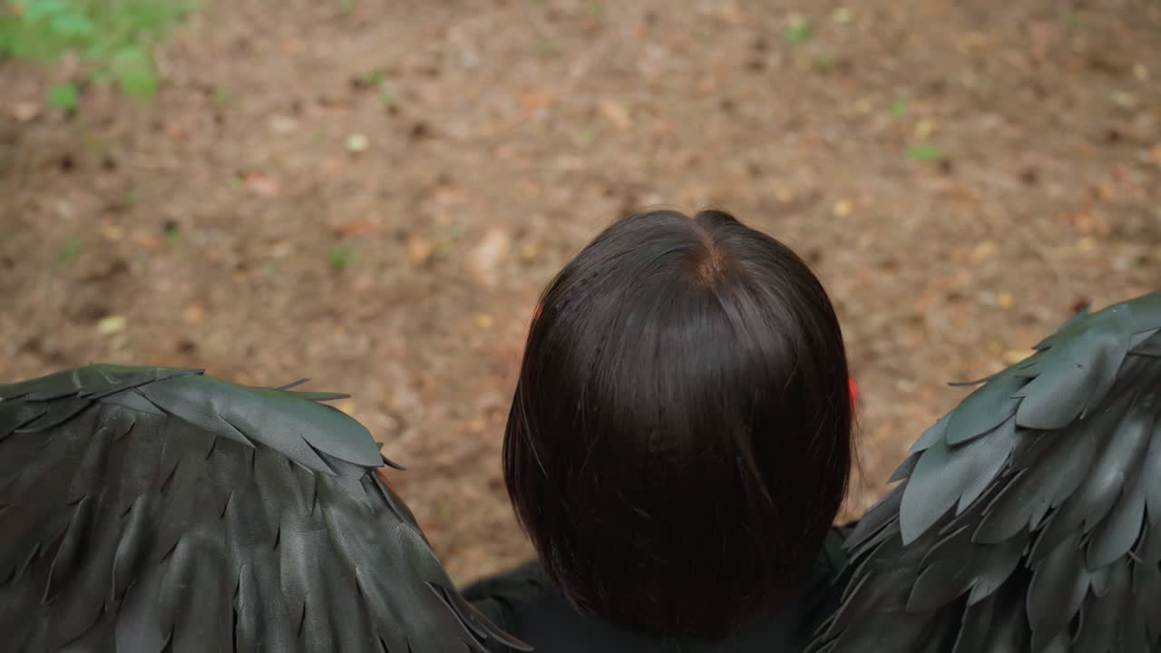 Aerial view of woman with black wings kneeling on forest floor surrounded by glowing red candles, meditating in calm spiritual ritual under soft woodland light filled with mystery