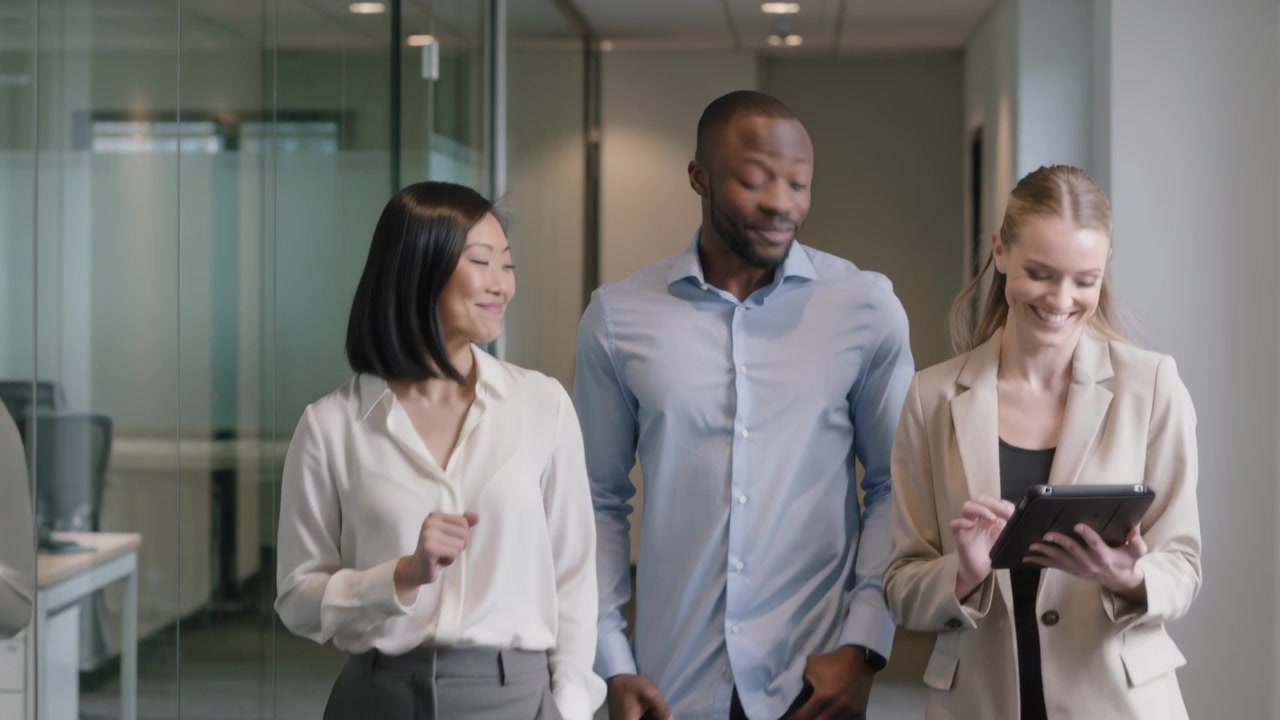 Entering frame, three colleagues walking toward camera sharing tablet in glass corridor, workwear