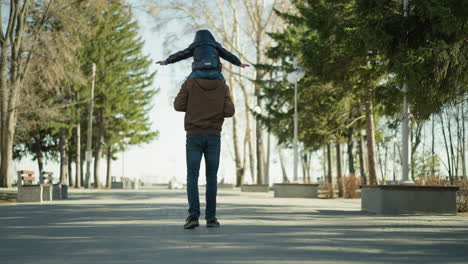 a father is seen carrying his son on his shoulders while walking down a path lined with trees and light poles, the child stretches his arms, seemingly scared of falling, as they move forward