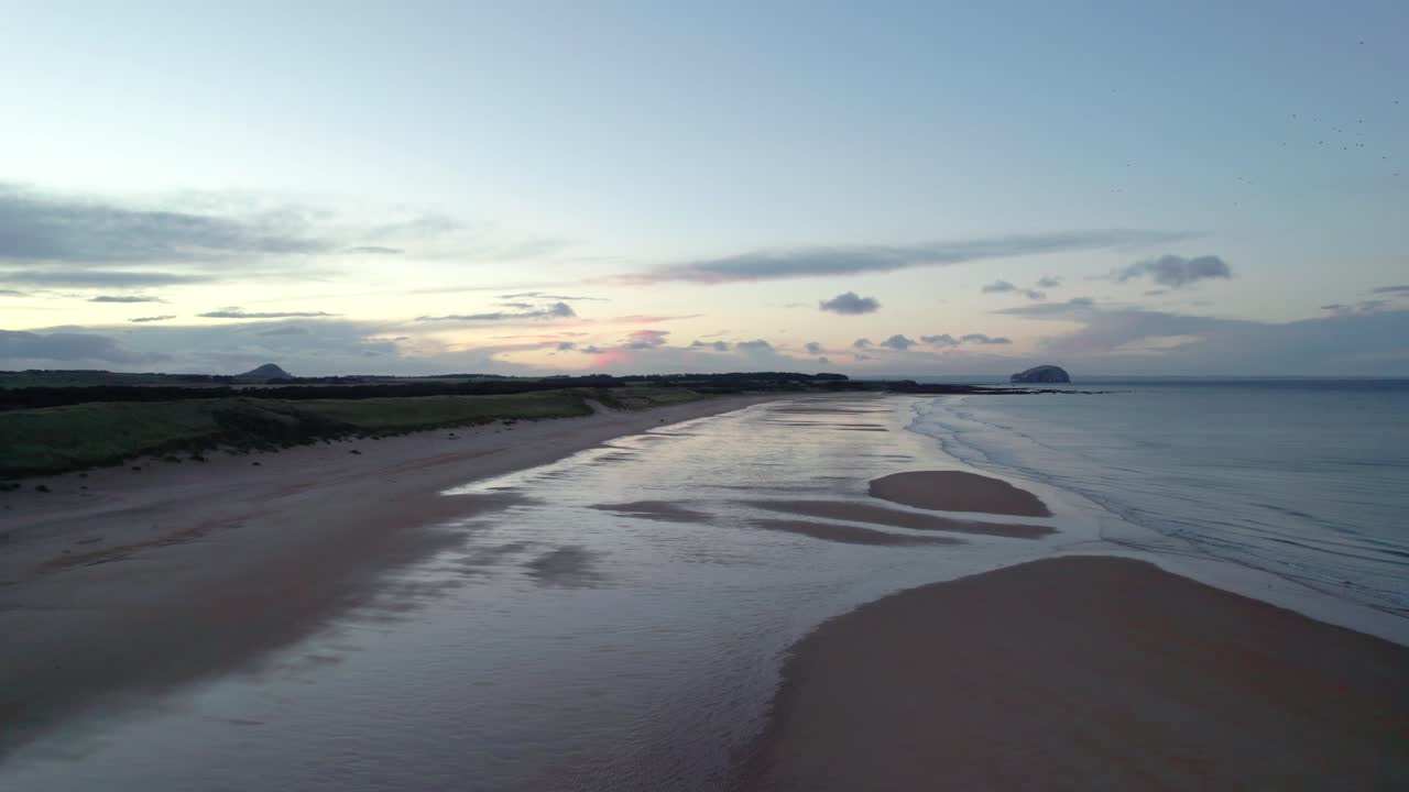 천천히 뒤로 날아가 긴 모래 해변 위로 떠오르는 드론 영상, 분홍색 일몰이 젖은 모래에 반사되고 조수가 해안을 부드럽게 감싸는 동안