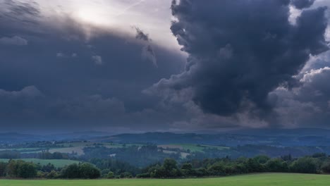 heavy stormy clouds roll above the summer landscape of hills, valleys, and forests with a distant village in the background