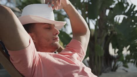 hombre biracial relajado sentado al sol en la playa poniendo sombrero sobre la cara, en cámara lenta