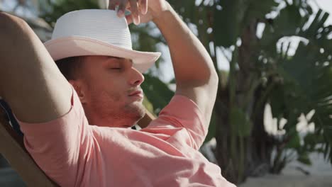 relaxed biracial man sitting in sun on beach putting hat over face, in slow motion