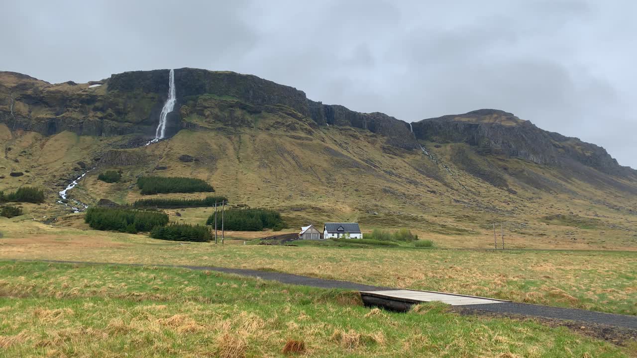 View over the high mountains around the tall waterfall Bjarnarfoss with a family home infront of it on a cloudy and misty day