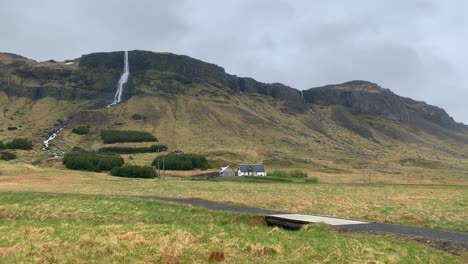 view over the high mountains around the tall waterfall bjarnarfoss with a family home infront of it on a cloudy and misty day