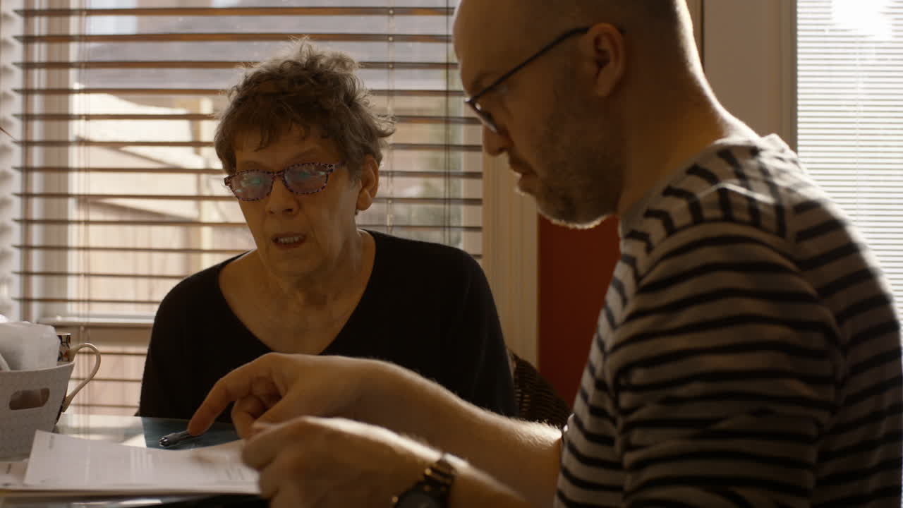 A senior woman sits at a kitchen table with a younger man. They are sorting, reviewing, and discussing documents together. Both wear reading glasses. Warmly backlit by the sun through window. 24fps.