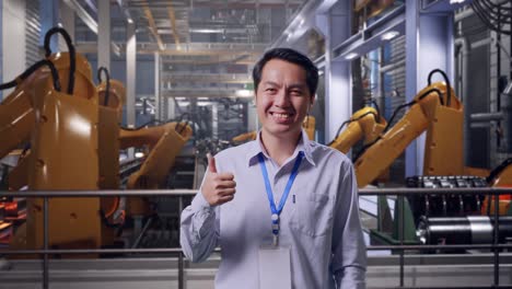 shot captures of an asian male professional worker standing with industrial robotic arms working, his broad smile at the camera and thumbs up
