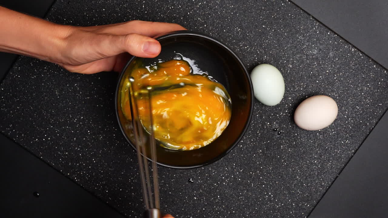 Munien sekoittaminen kulhossa, jossa on eri värejä tummalla keittiön pinnalla. Käsi sekoittaa keltaisia munia kulhossa, ja kaksi munaa lepää lähellä tummalla pinnalla.