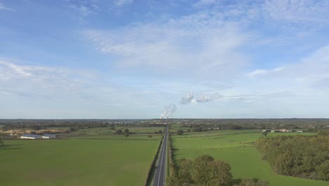 Cooling-Towers-Emitting-Smoke-At-Industrial-Thermal-Power-Station-Seen-From-Long-Straight-Road-In-Countryside