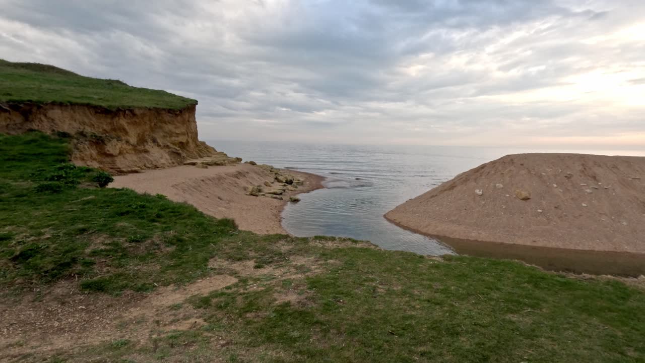 Video moving forward towards an ocean inlet river on a Jurassic South Coast beach. This stabilised footage starts on grass then moves forward to reveal the river cutting through the sand on cloudy day